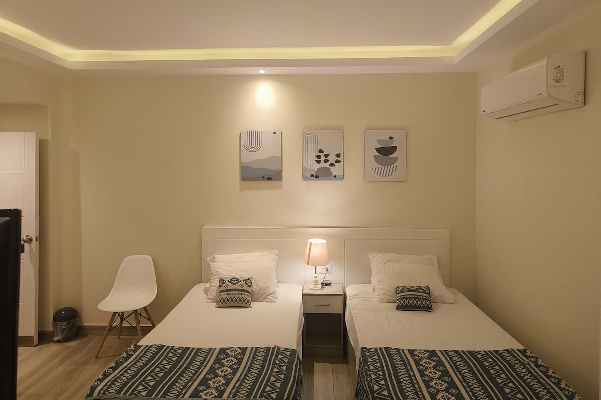 A hotel room with two beds, a bedside table with a lamp, a chair, and wall art.