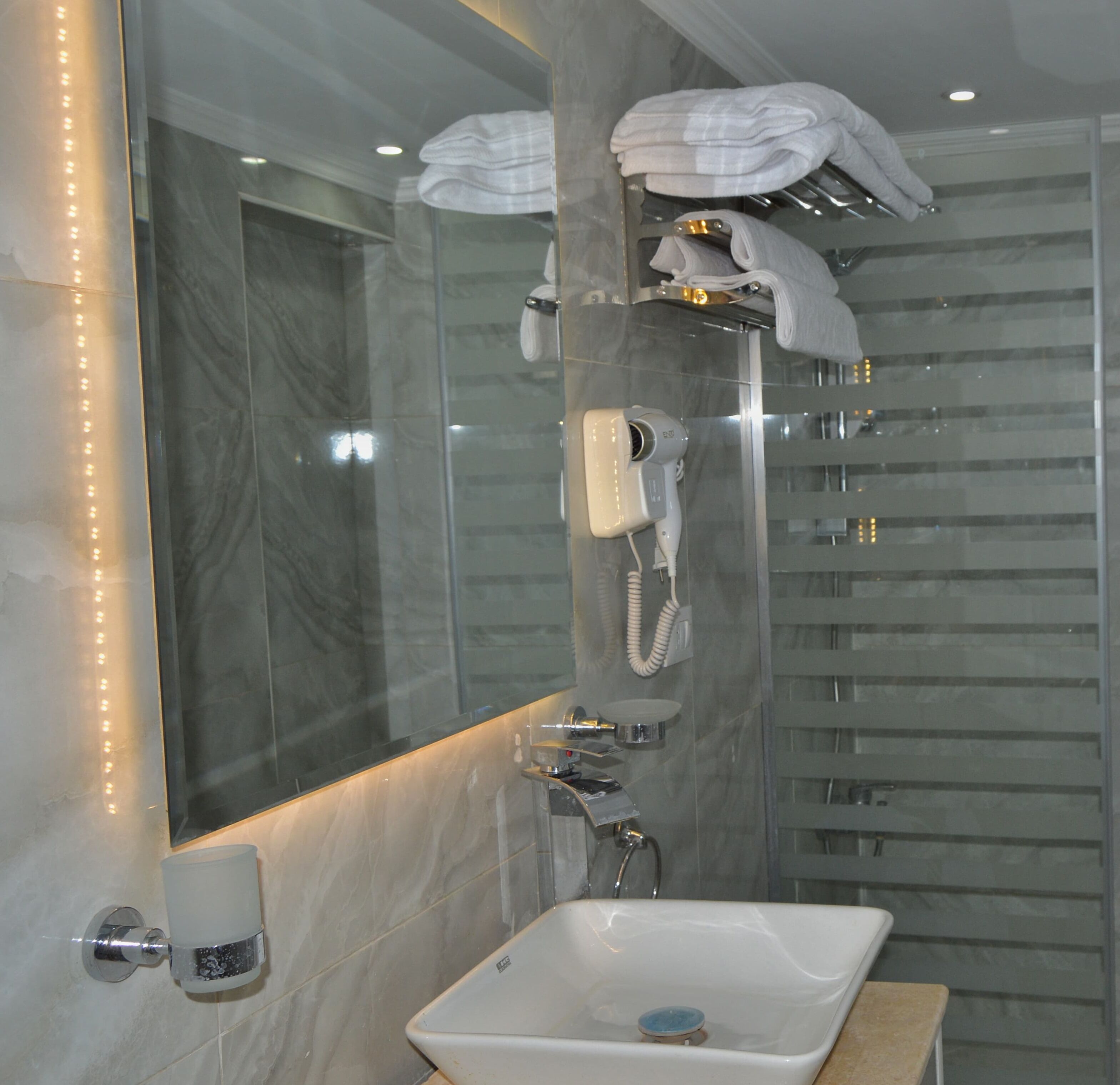 A modern bathroom with a marble wall, a large mirror, a sink, a handheld showerhead, and a towel rack with folded towels.