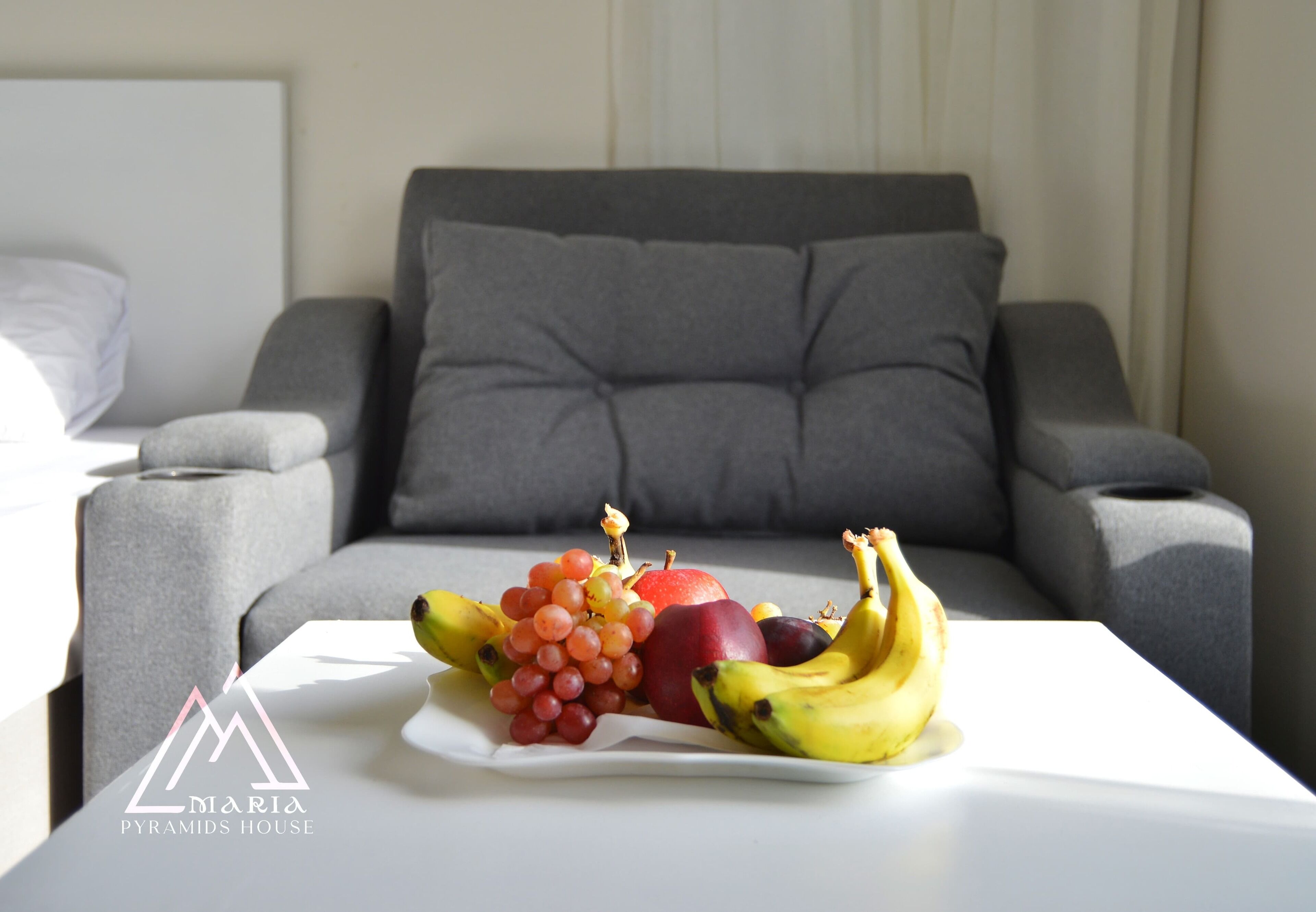 A fruit platter on a white table in a modern living room with a grey sofa and a white headboard.