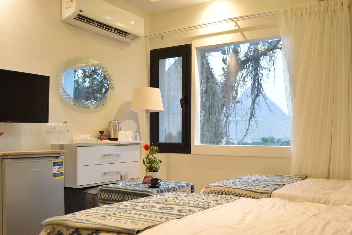 A hotel room with two beds, a desk with a computer, a small fridge, a window with a view of trees, and a wall-mounted air conditioner.