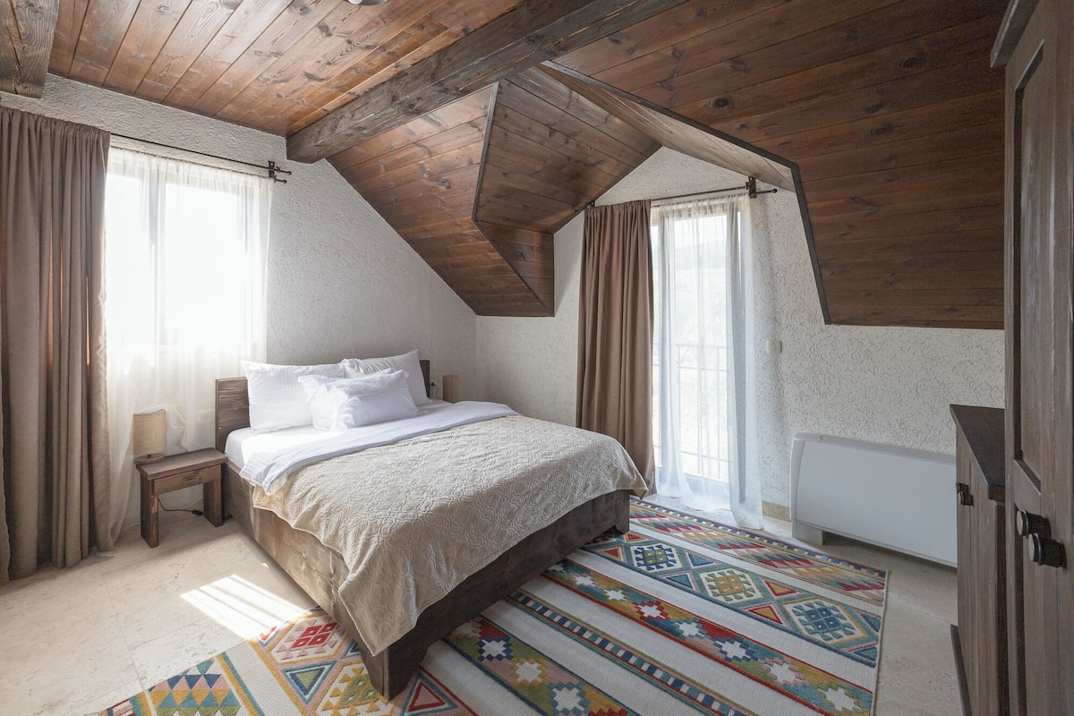 A bedroom with a sloped ceiling, a bed with white linens, a bench, and a colorful rug.