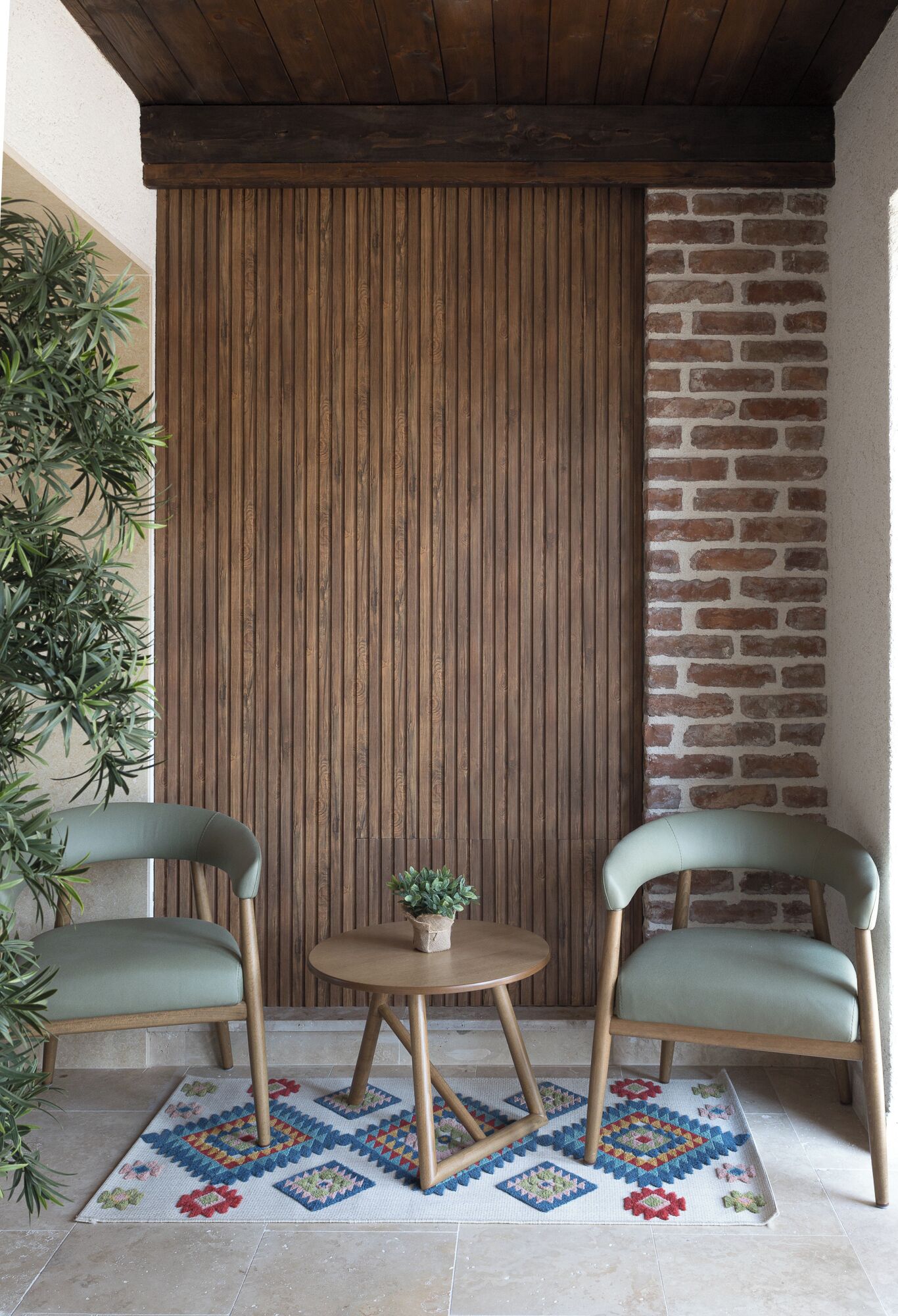 A small, cozy seating area with two chairs and a table, a wooden slatted wall, and a brick wall.