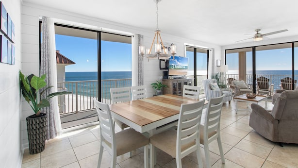 A spacious living area with a wooden dining table, white chairs, a sofa, and a TV. A balcony with a view of the ocean is visible.
