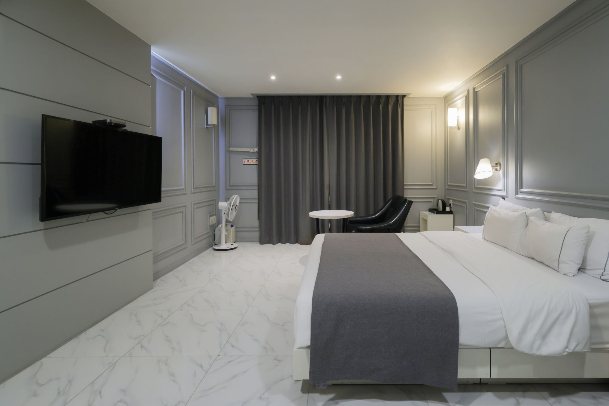 A modern hotel room with a large bed, a flat-screen TV mounted on the wall, a small round table, a chair, and a bedside lamp.