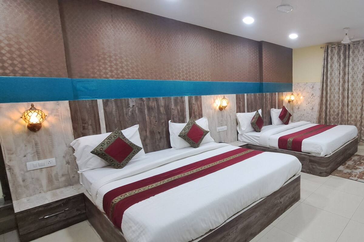 A hotel room with two beds, each with white bedding and red and gold decorative pillows. The room features wooden headboards, a blue accent wall, and a patterned curtain.