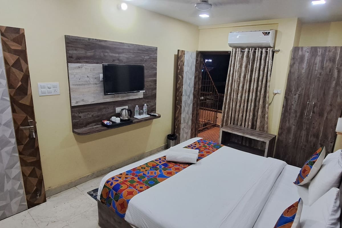 A hotel room with a bed, a TV mounted on the wall, a small table with a kettle and cups, and a curtain.