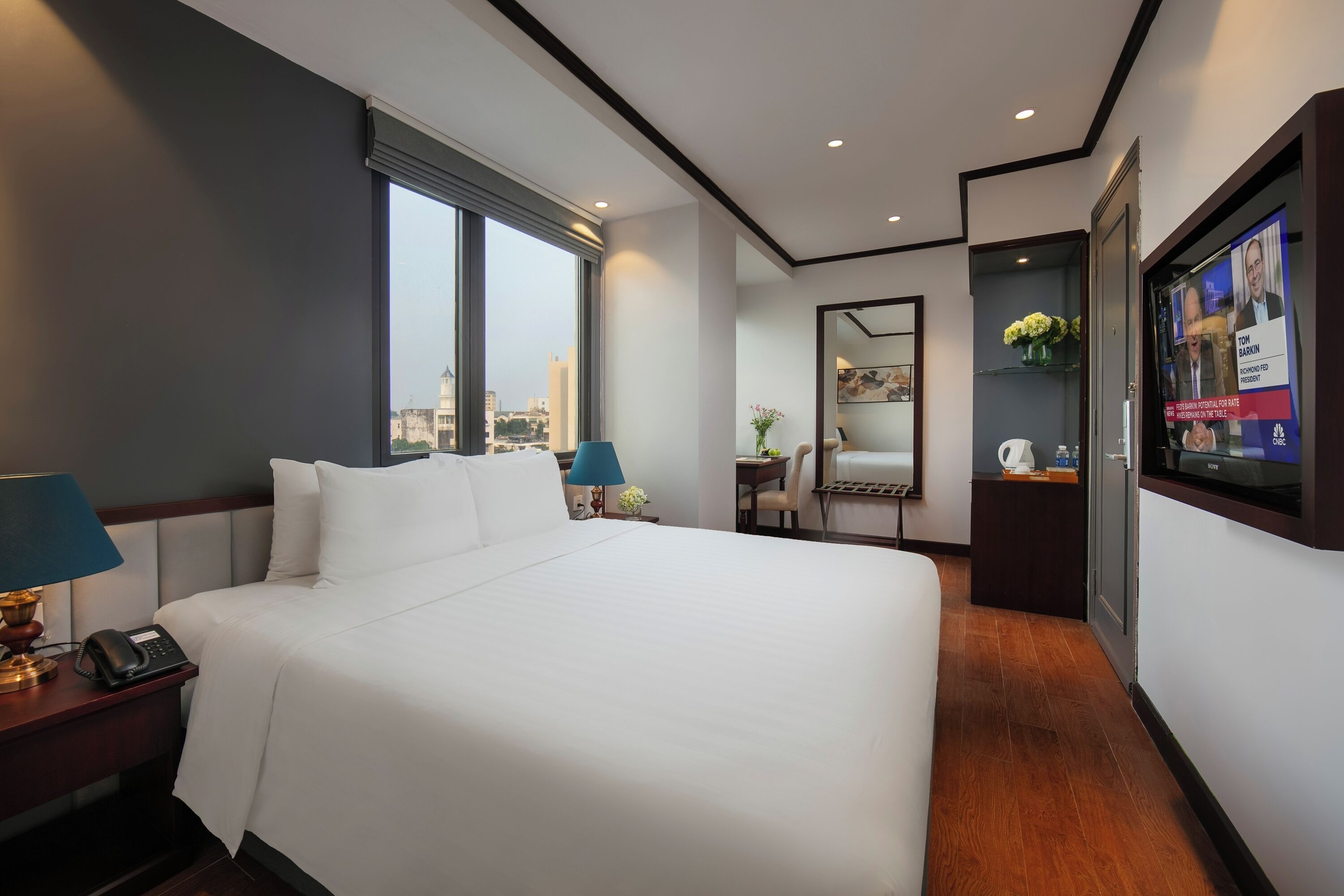 A hotel room with a large bed, a desk with a computer, a TV, and a window with a city view.