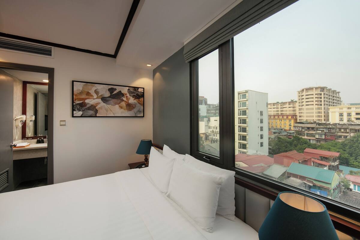 A hotel room with a large bed, a painting on the wall, a nightstand with a lamp, and a window with a city view.