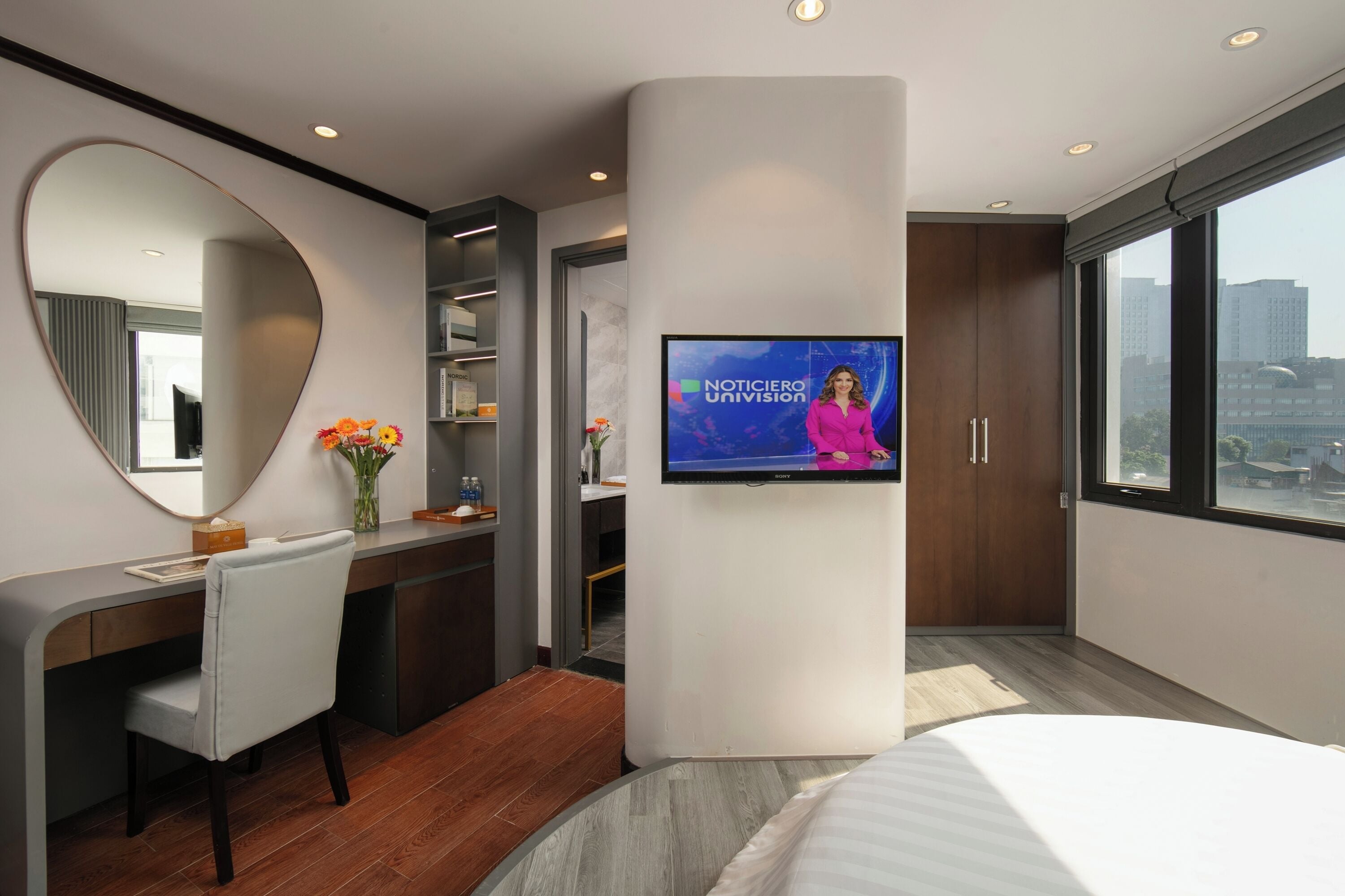 A modern hotel room with a large TV, a desk with a chair, a bed, and a large window offering a city view.