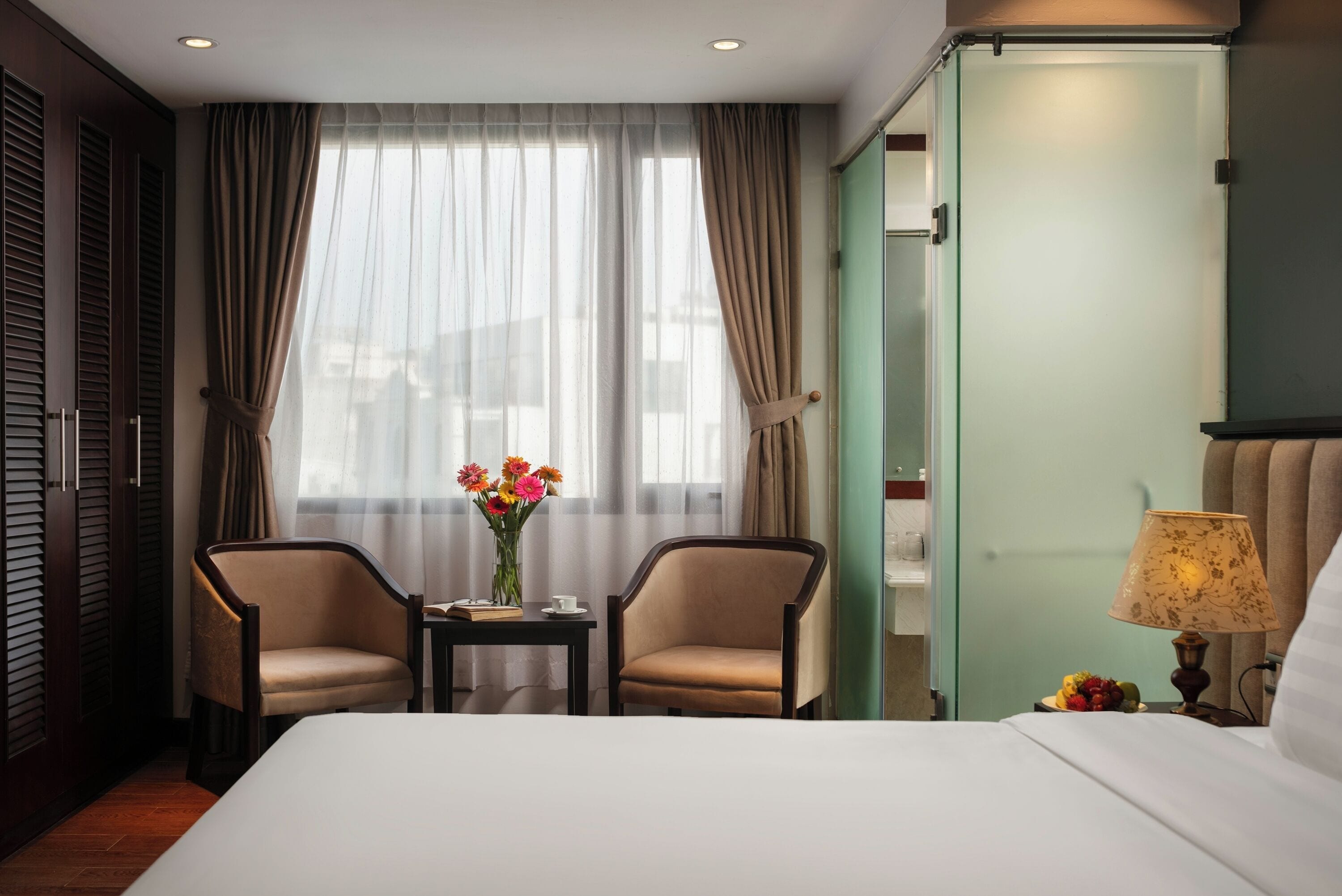 A hotel room with a bed, two armchairs, a small table with a vase of flowers, a lamp, and a view of the outside through sheer curtains.
