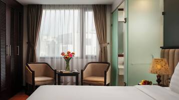 A hotel room with a bed, two armchairs, a small table with a vase of flowers, a lamp, and a view of the outside through sheer curtains.