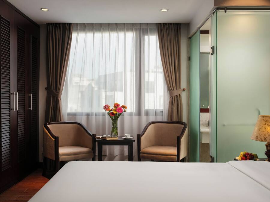 A hotel room with a bed, two armchairs, a small table with a vase of flowers, a lamp, and a view of the outside through sheer curtains.