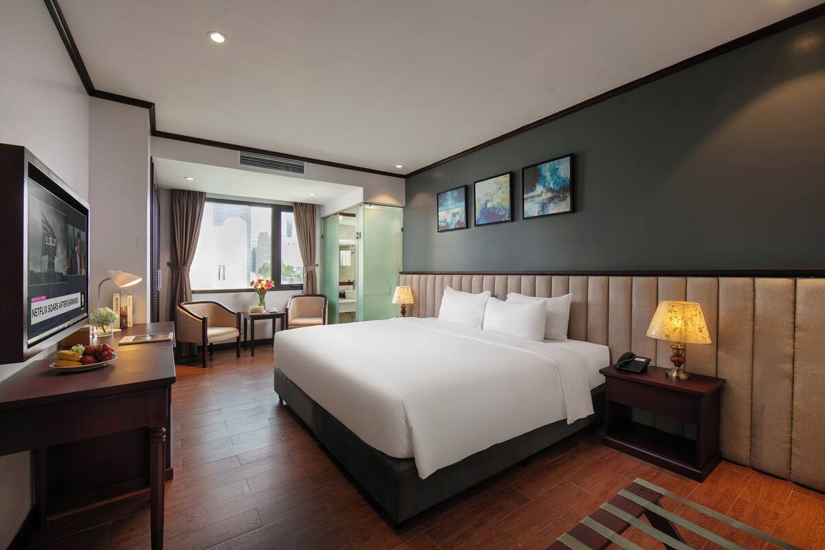 A hotel room with a large bed, bedside table, TV, and a desk with a chair.