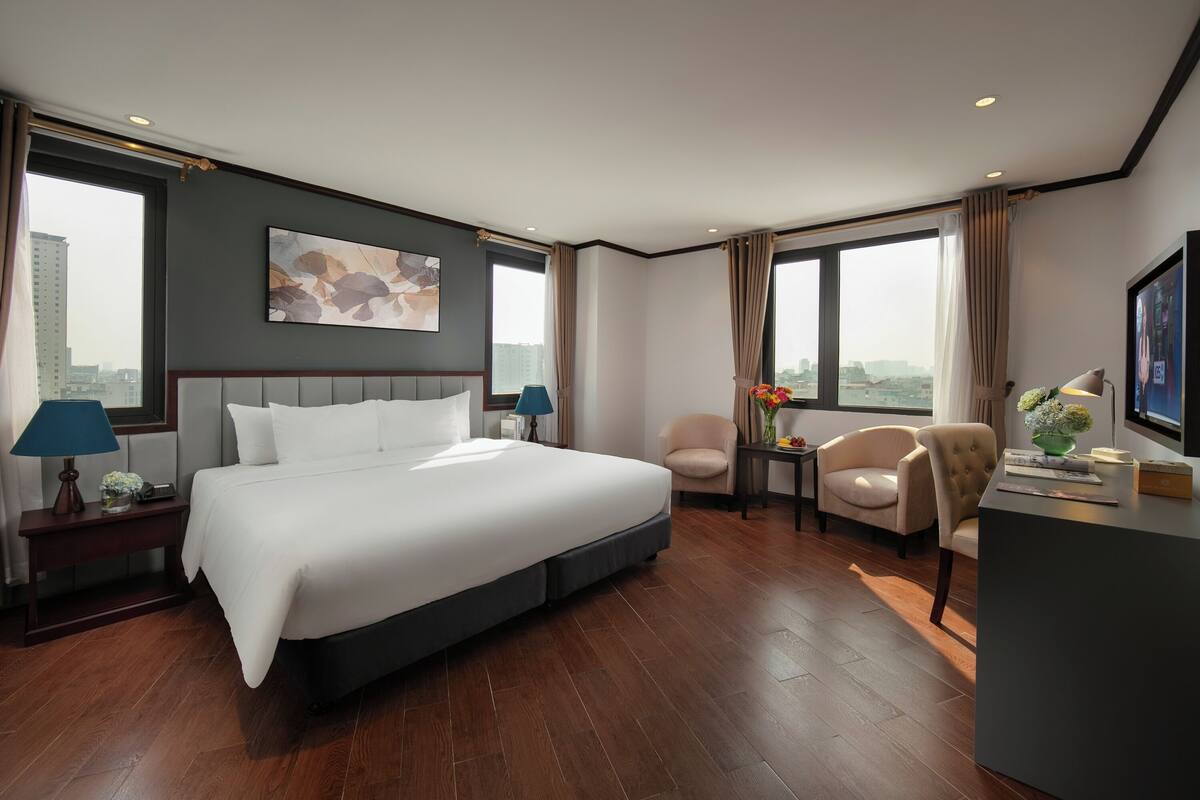 A modern hotel room with a large bed, two armchairs, a desk, and a view of the city.