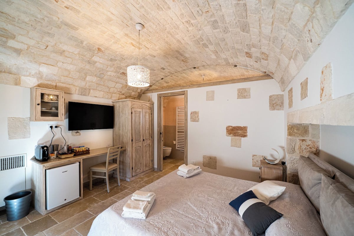 A bedroom with a stone ceiling, a bed with a patterned duvet, a desk with a computer, a wardrobe, and a television on the wall.