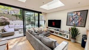A modern living room with a grey sectional sofa, a flat-screen TV, and a large window overlooking a garden.