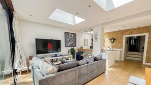 A modern living room with a grey sofa, a flat-screen TV, and a kitchen area with a brick wall.