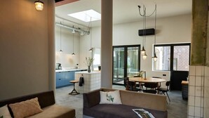 En moderne stue med sofa og spisebord, et køkken i baggrunden og store vinduer.