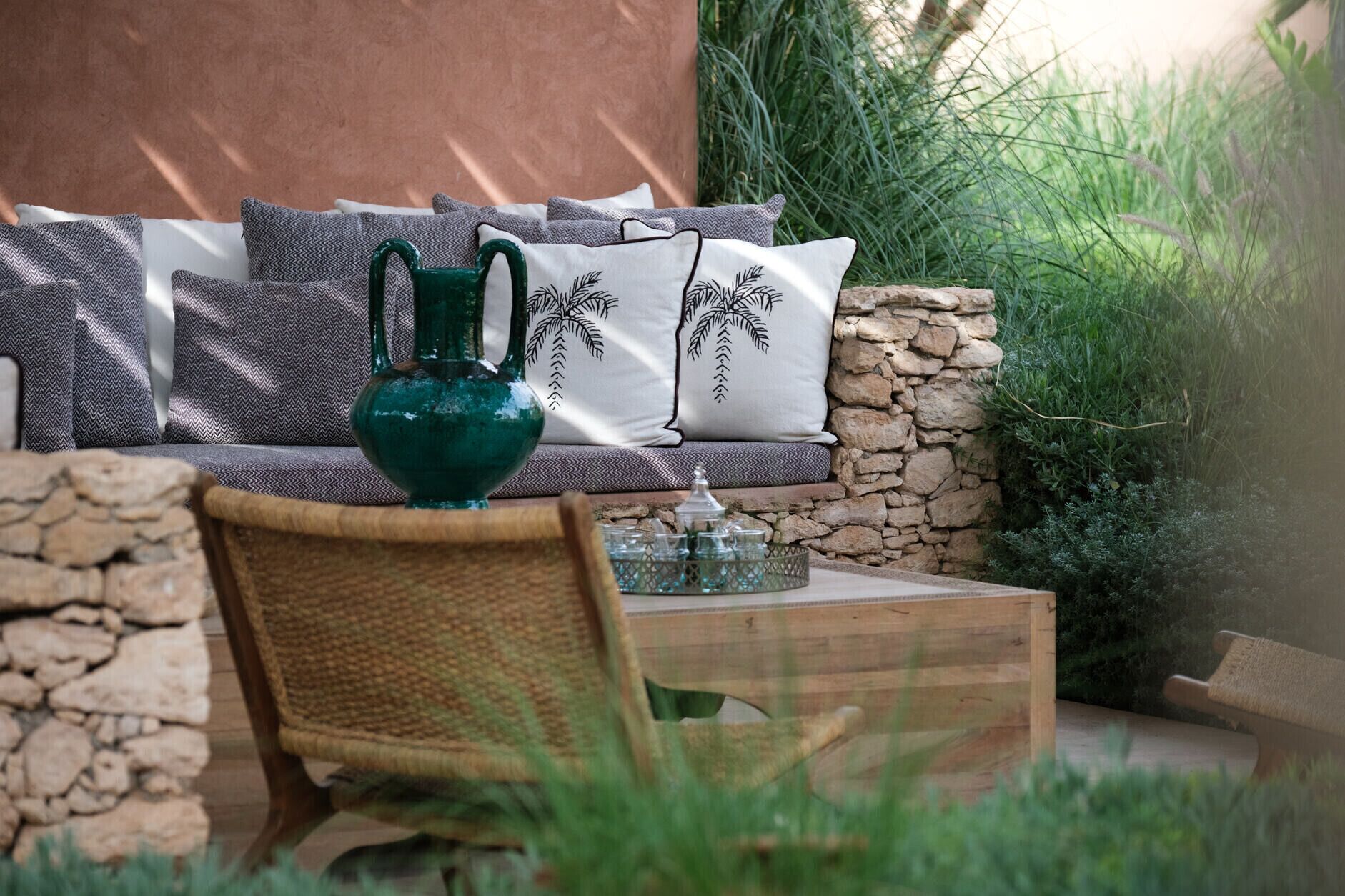 Un muro de piedra, una silla de mimbre, una mesa de madera y un sofá con cojines estampados y un jarrón verde.