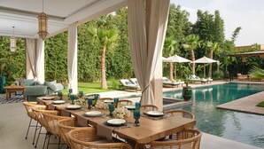 An outdoor dining area with wicker chairs and tables, a pool, and palm trees in the background. - NETUMIA (Marrakech)