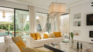 A modern living room with a white sofa, yellow cushions, a glass coffee table, and a large window overlooking a pool and greenery. - NETUMIA (Marrakech)