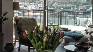 Ein Wohnzimmer mit einem großen Fenster und Blick auf eine Küstenstadt, ein Korbstuhl, eine Vase mit Blumen und ein Sofa mit einem Fellüberwurf.