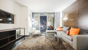 A modern living room with a flat-screen TV, a glass coffee table, a grey sofa with orange cushions, and a large window offering a city view. - SVS: Strip View Studio | Open Balcony | Full Size Fridge | Palms Place (Las Vegas)