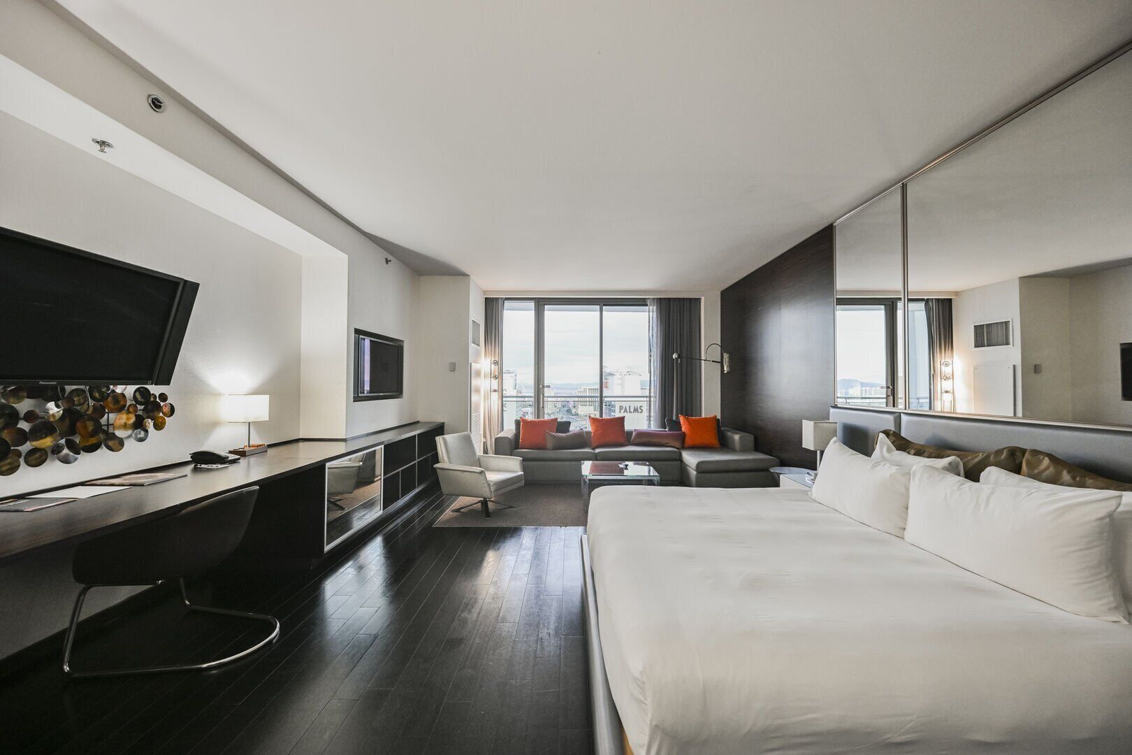 A modern hotel room with a large bed, a desk with a flat-screen TV, a seating area with a sofa and chairs, and large windows offering a city view.