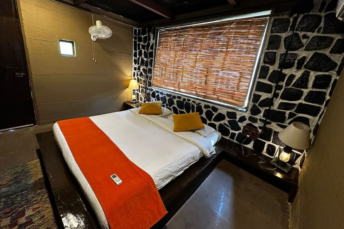 Ein Hotelzimmer mit einem Bett, einer Steinwand, einem Fenster mit Jalousien, einem Nachttisch mit Lampe und einem kleinen Tisch ebenfalls mit einer Lampe.