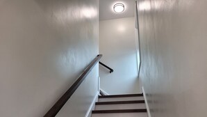 A staircase with wooden steps and a handrail, leading upwards in a narrow hallway with white walls and a ceiling light. - The Traveler on Main Suite #12- Across the Street from Back of the Dragon! (Tazewell)