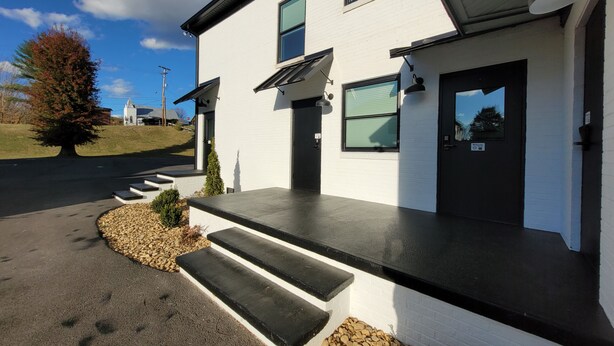 A modern two-story building with a white exterior, black trim, and a black roof. There are steps leading to the front door, a small garden area with a tree, and a clear sky in the background. - The Traveler on Main Suite #12- Across the Street from Back of the Dragon! (Tazewell)