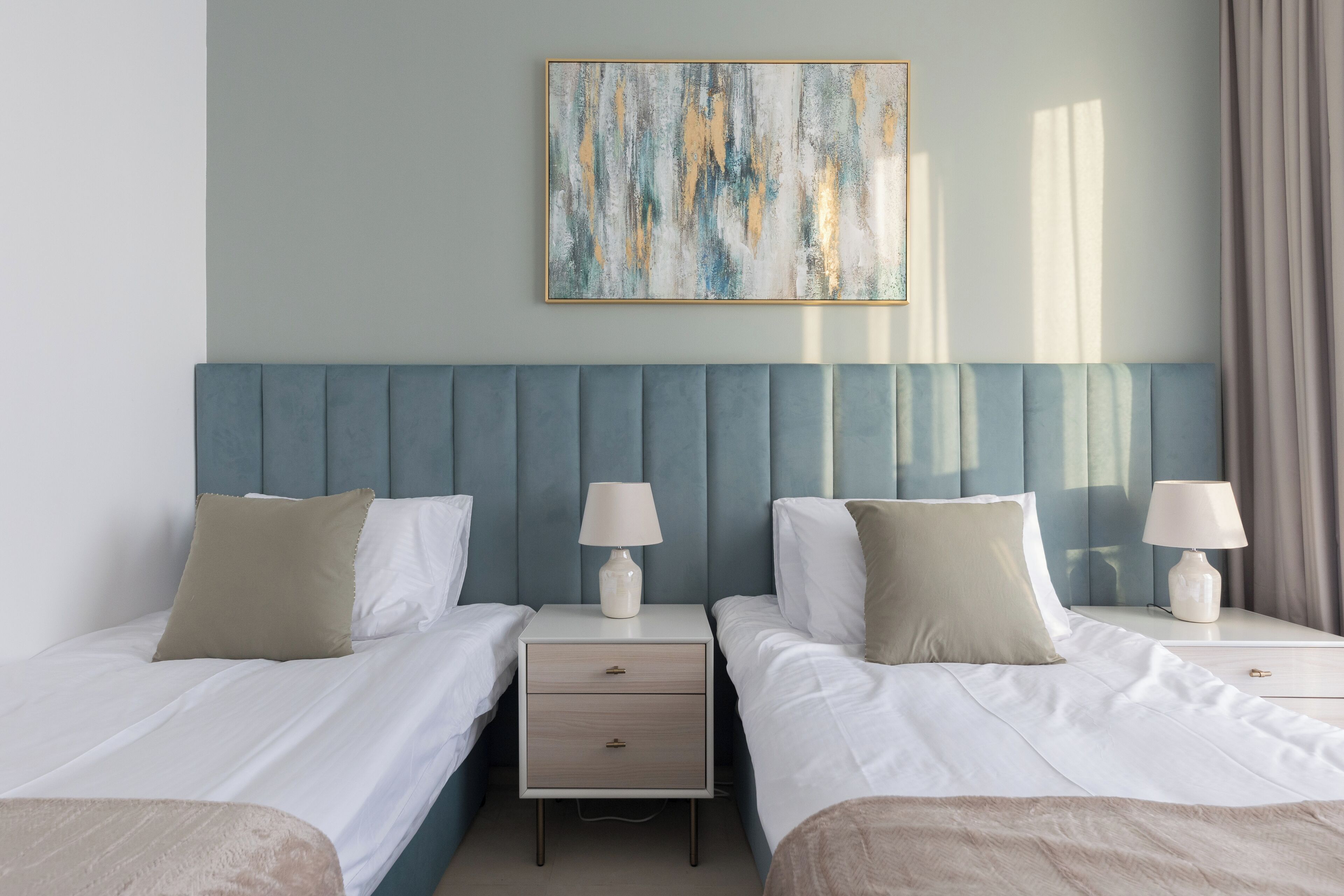 A hotel room with two beds, a headboard, bedside tables with lamps, a small drawer, and a painting on the wall.
