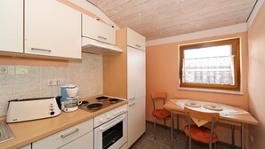 A compact kitchen with white cabinets, a stove, a coffee maker, and a window with a curtain. - Villa Hügl - cozy vacation apartment for couples (Waldkirchen)