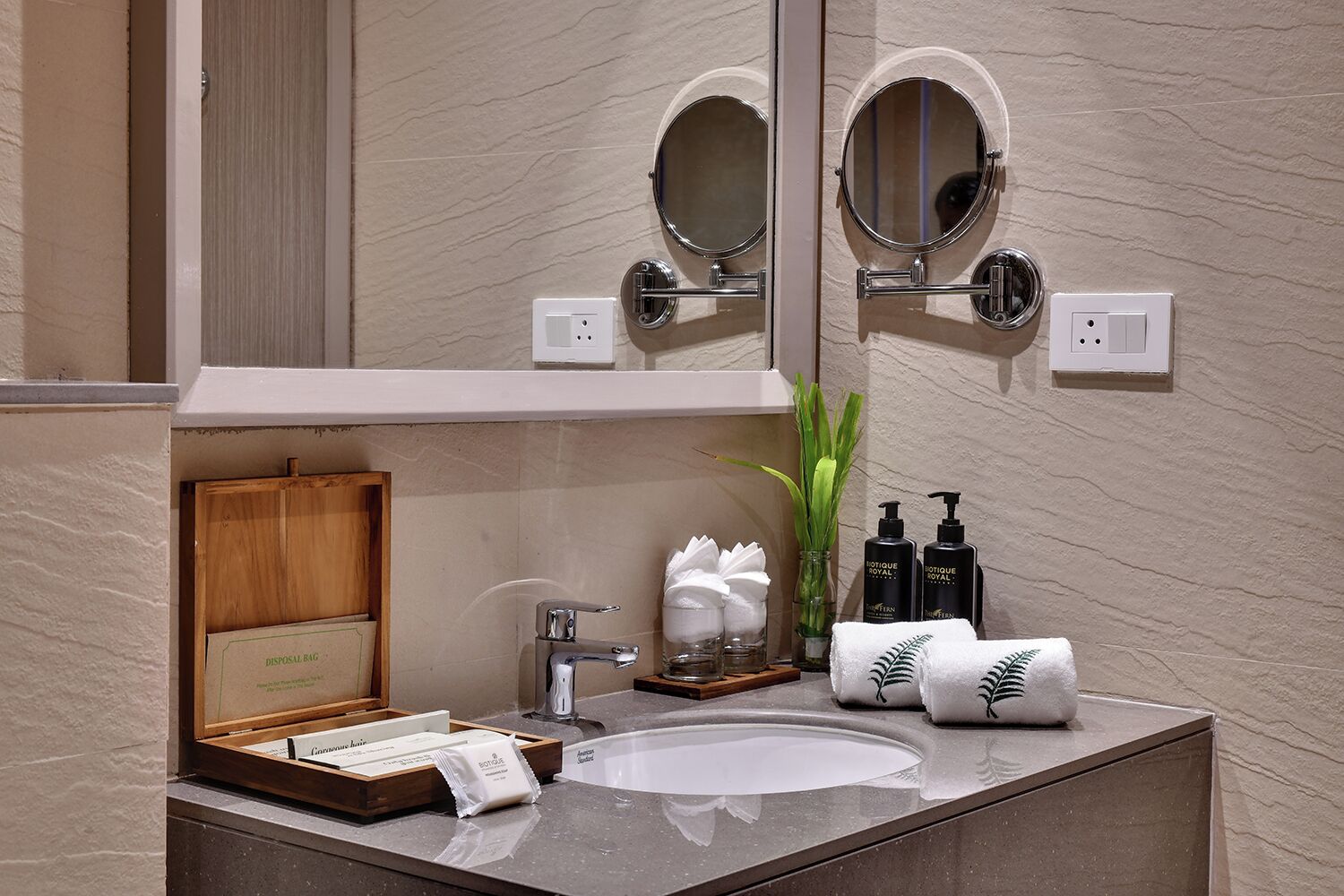 Un baño moderno con lavamanos de mármol, dos espejos redondos, un mueble de tocador con artículos de aseo personal y un estuche de madera con la etiqueta 'DISPOSAL BAG'.