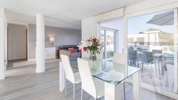 A modern dining area with a glass table and chairs, a sofa in the background, and large windows offering a view of the outdoors.