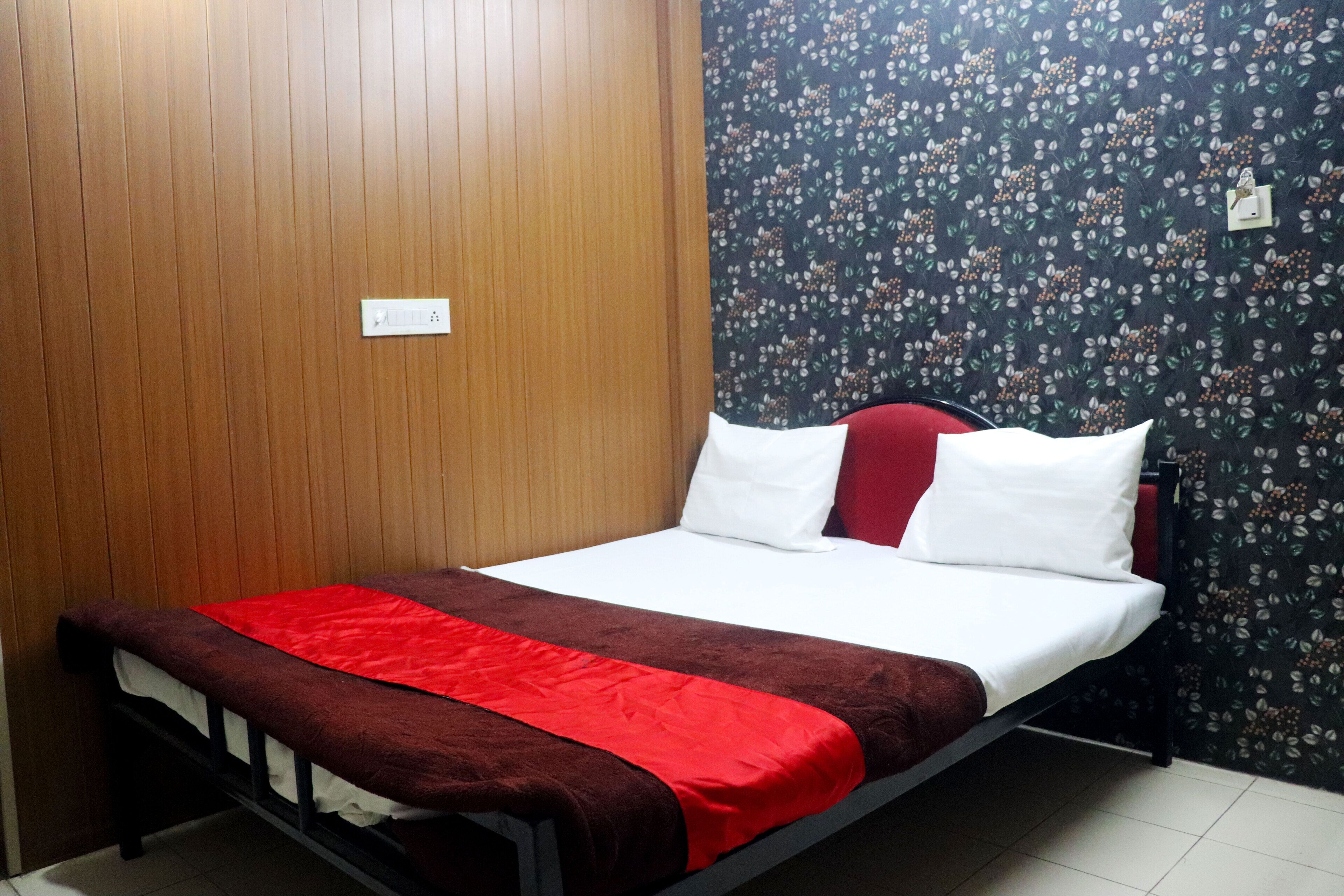 A single bed with a red and white bedspread, a wooden headboard, and a floral-patterned wall.