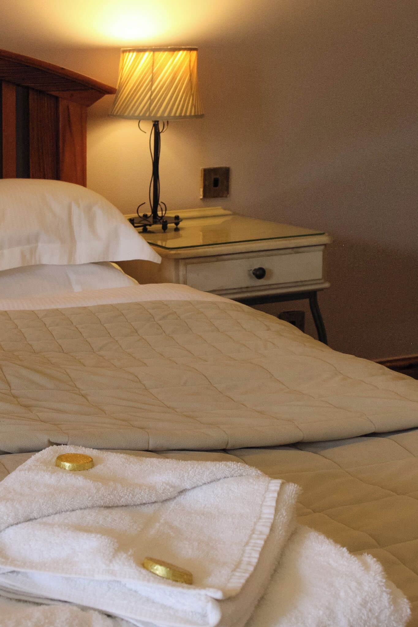 A neatly made bed with white linens, a bedside lamp, and a small bedside table with a drawer.