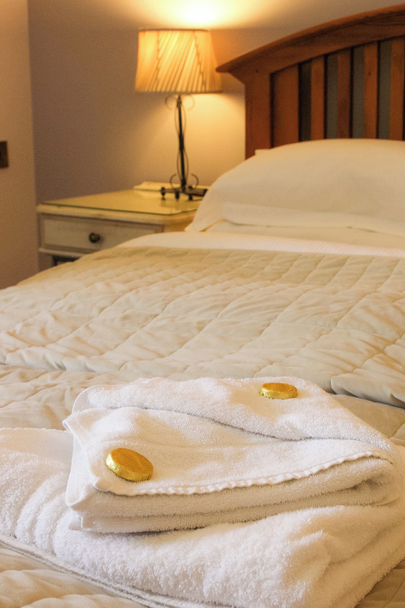 A neatly made bed with a quilted white duvet, a folded white towel with a gold tag, and a bedside lamp on a wooden nightstand.