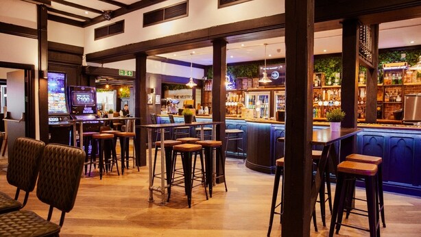 A bar area with stools, a bar counter, and a television screen displaying a game. - The Vestry (Chichester)