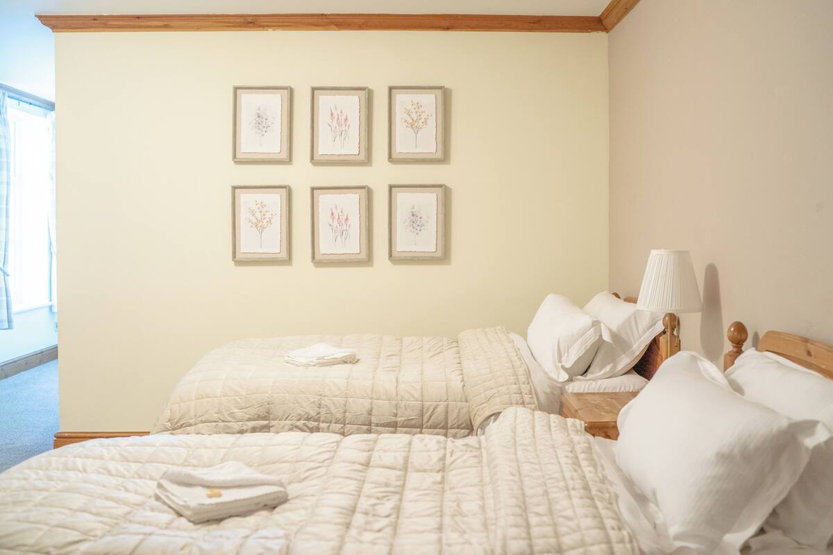 Habitación de hotel con dos camas, cabecero de madera, mesita de noche con lámpara y cuadros de motivos botánicos en la pared.