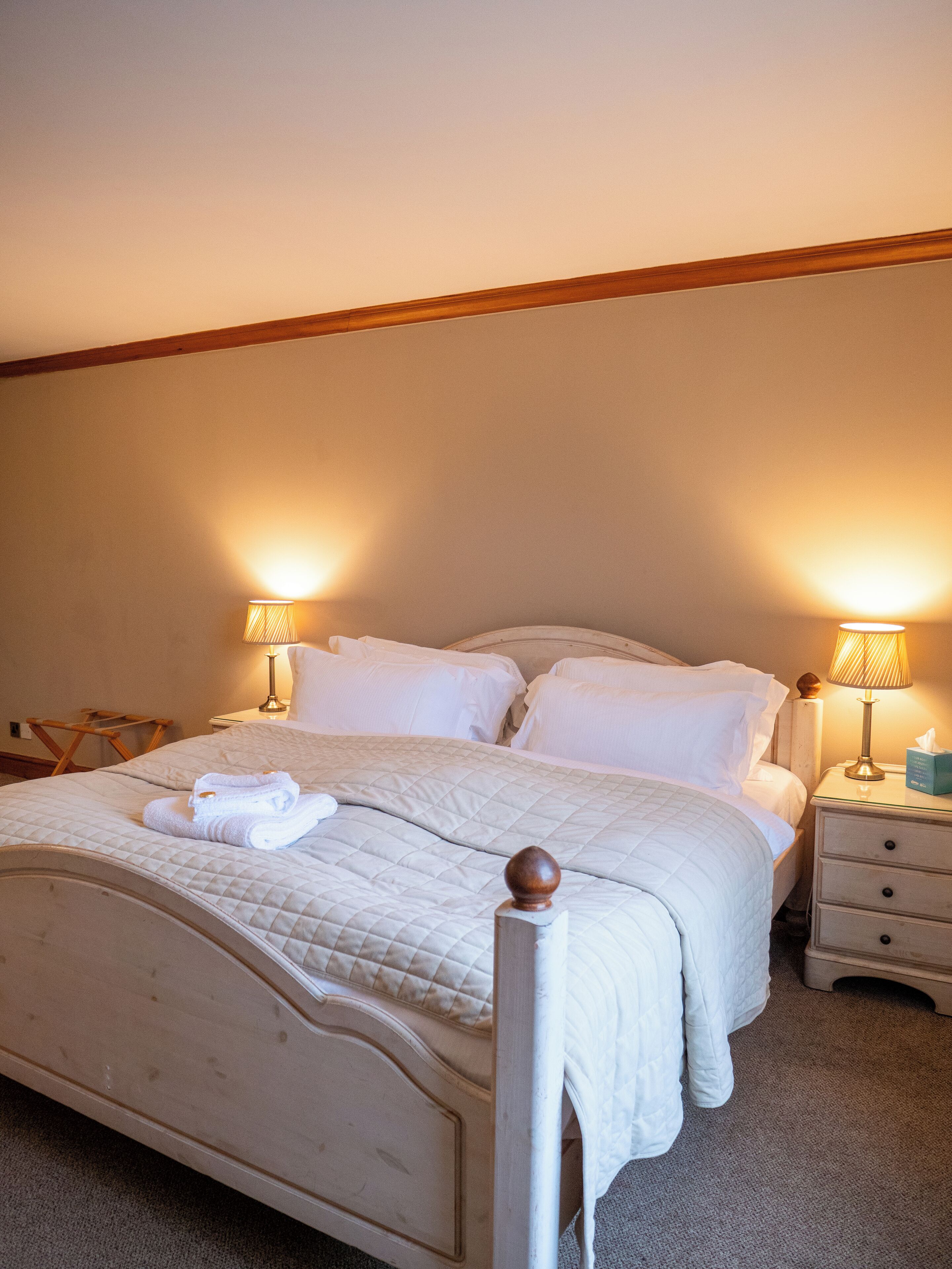 A neatly made bed with white linens and a folded towel, two bedside lamps, a wooden headboard, and a nightstand with a tissue box.