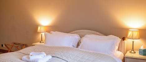 Una cama bien hecha con sábanas blancas y una toalla doblada, dos lámparas de noche, un cabecero de madera y una mesita de noche con un dispensador de pañuelos.