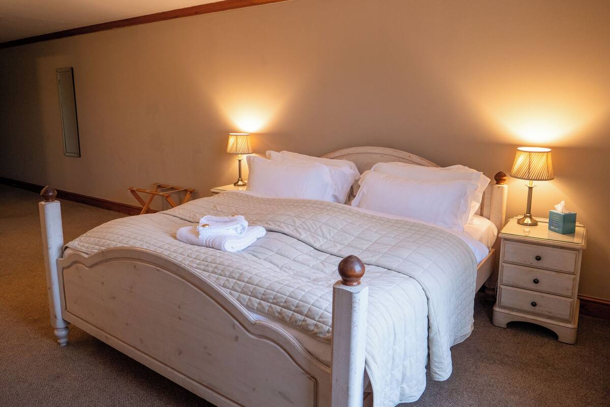 Una cama bien hecha con sábanas y almohadas blancas, dos lámparas de noche, una mesita de noche con una caja de pañuelos y una silla de madera.
