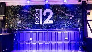 A stage with a backdrop labeled "ROOM 12," surrounded by sound equipment and a wall decorated with plants. - The Vestry (Chichester)