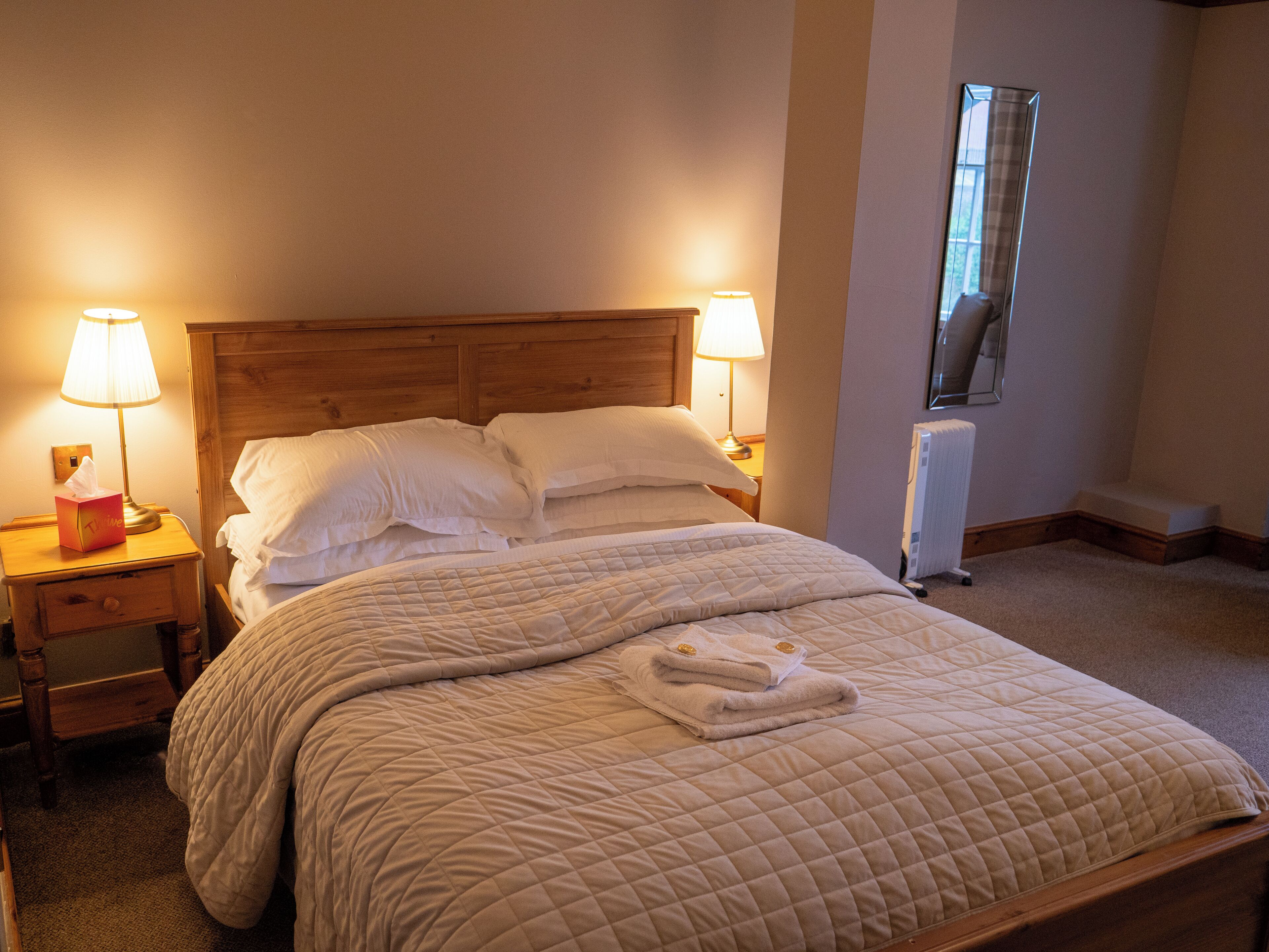 A neatly made bed with a quilted blanket, two bedside lamps, a nightstand with a tissue box, and a radiator.