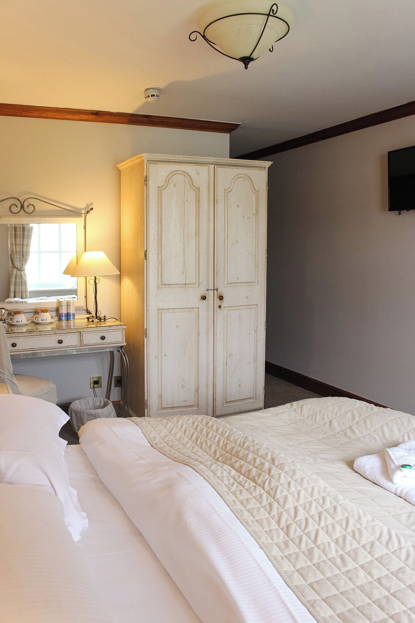 A hotel room with a bed, a desk with a lamp, a wardrobe, and a television on the wall.