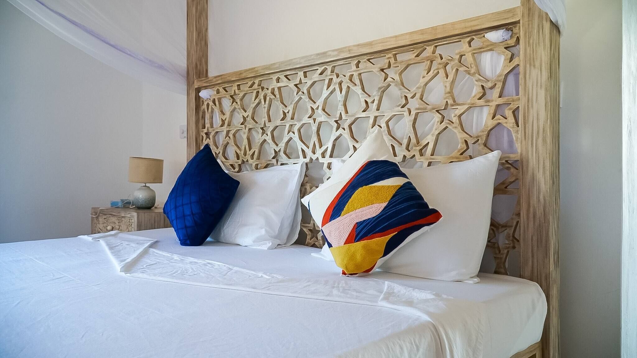 A bed with white linens, a wooden headboard with intricate carvings, and two pillows in blue and multicolored patterns.