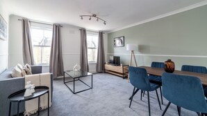 A modern living room with a sofa, a glass coffee table, a dining table with chairs, a TV on a stand, and a lamp. - The Peckham Luxury Apartment (London)