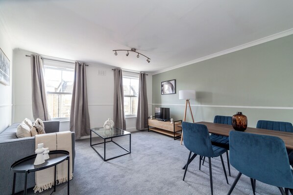 Un salon moderne comprenant un canapé, une table basse en verre, une table à manger avec des chaises, un téléviseur posé sur un meuble et une lampe.