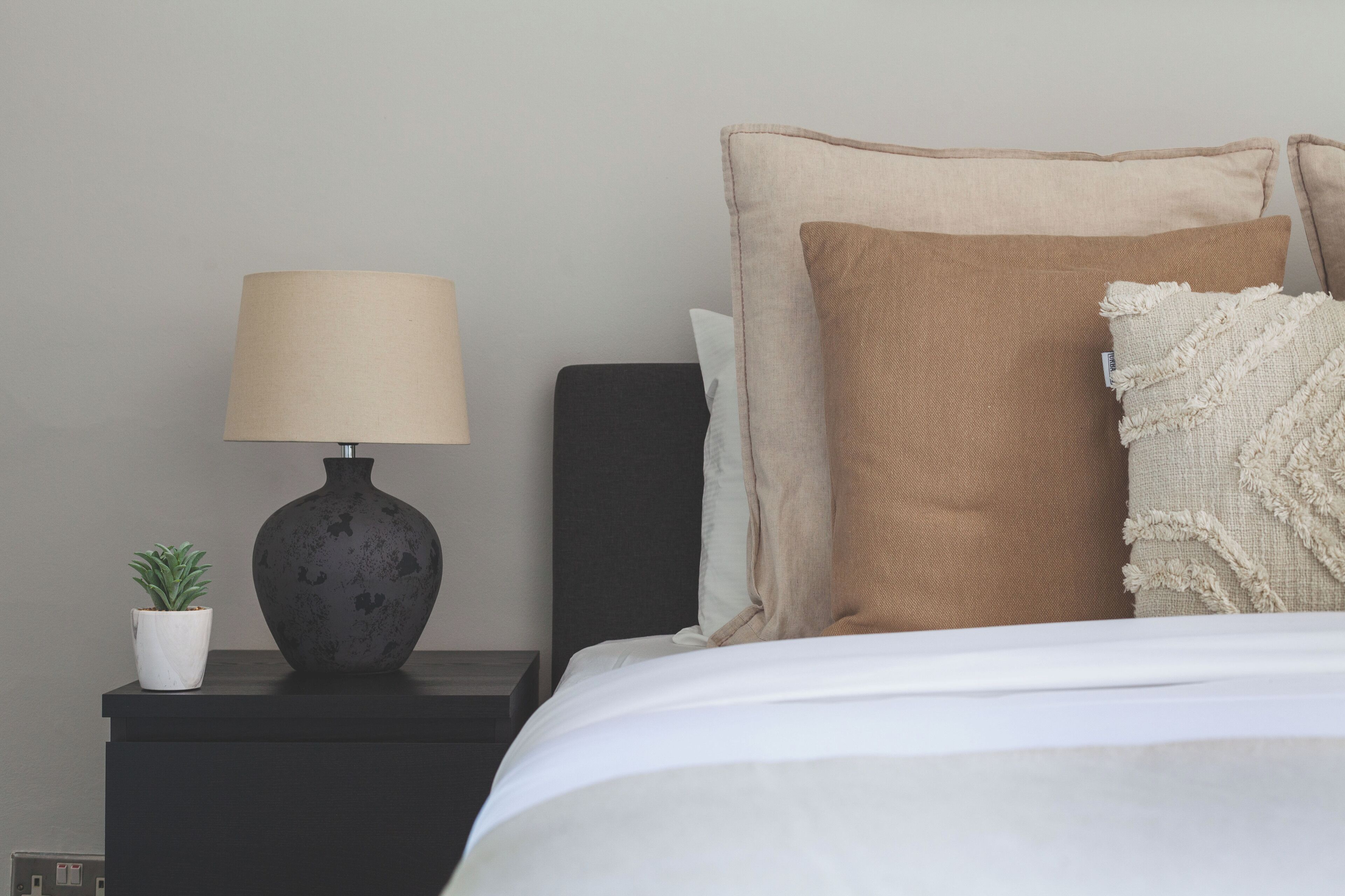 Ein ordentlich bezogenes Bett mit weißer Decke und mehreren Kissen, ein dunkler Nachttisch mit einer Lampe und einer kleinen Topfpflanze, sowie eine schlichte Wand im Hintergrund.